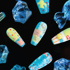 3D press-on nails from the "Candy Night" collection, featuring coffin-shaped nails with vibrant, whimsical designs in pastel yellows, blues, and glitter—including hearts, dots, and text ("LOVE IN PARK"). The nails are displayed against a black background with surrounding blue, faceted crystal-like decorations, emphasizing their playful, candy-inspired aesthetic.