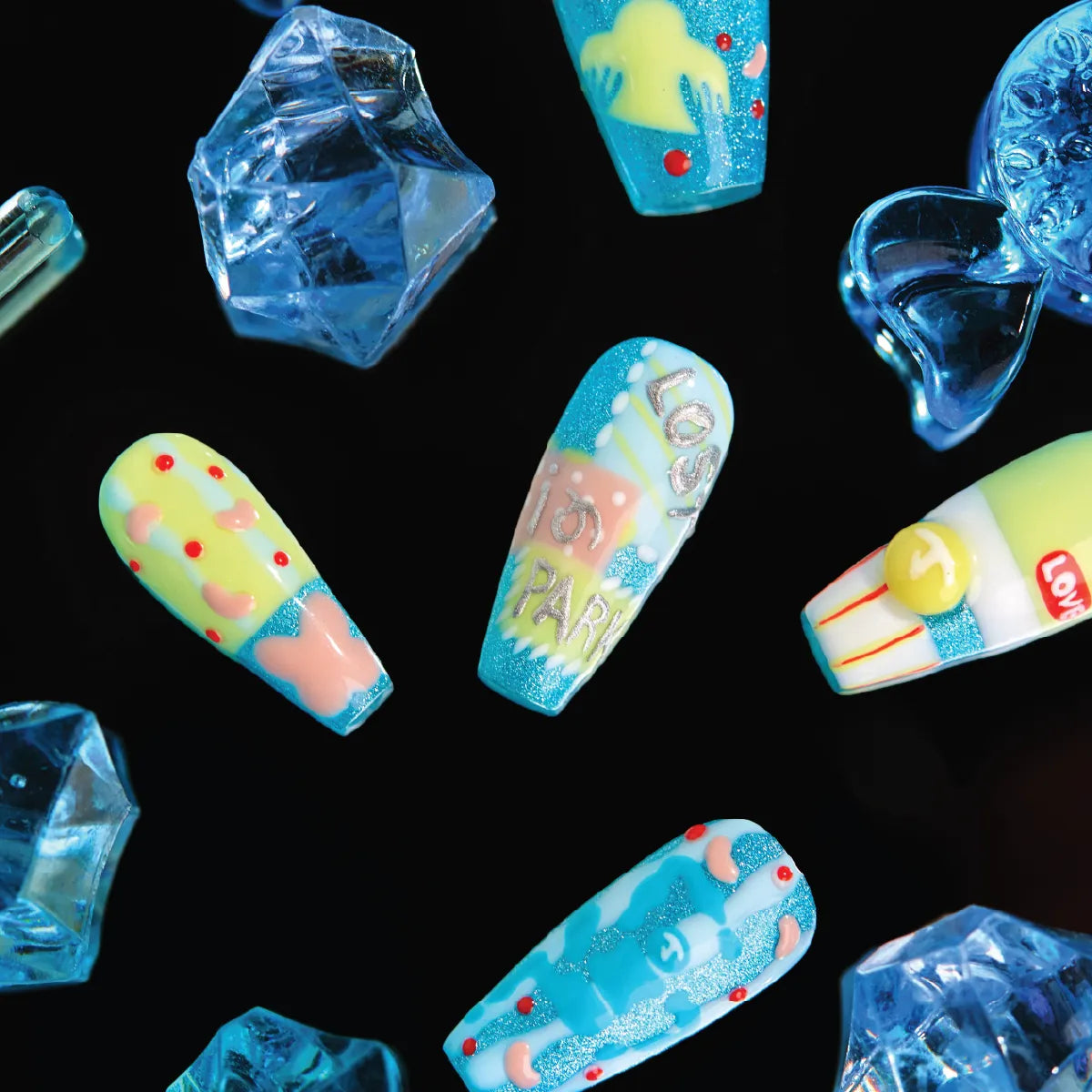 3D press-on nails from the "Candy Night" collection, featuring coffin-shaped nails with vibrant, whimsical designs in pastel yellows, blues, and glitter—including hearts, dots, and text ("LOVE IN PARK"). The nails are displayed against a black background with surrounding blue, faceted crystal-like decorations, emphasizing their playful, candy-inspired aesthetic.