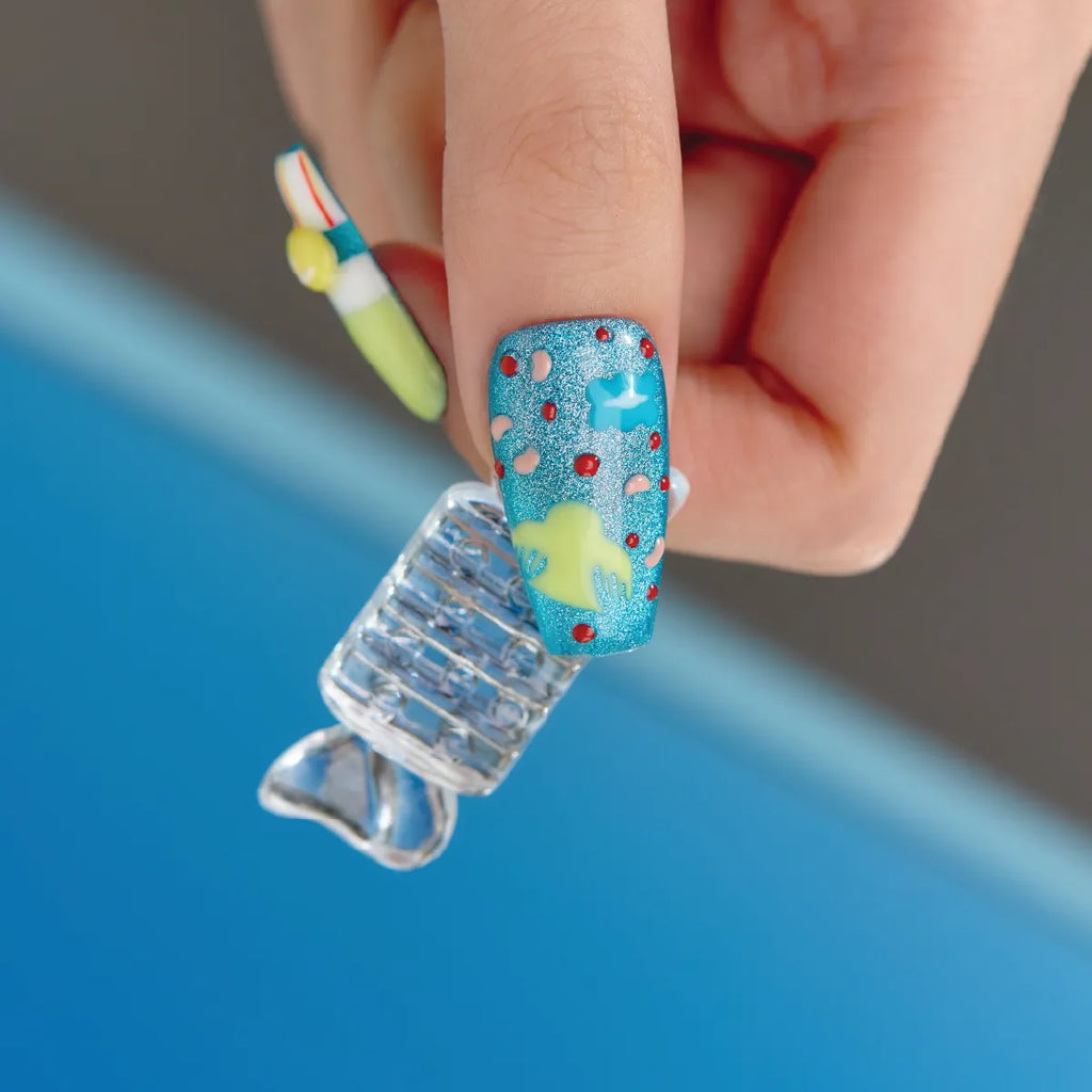 Close-up of a hand holding a 3D press-on coffin nail from the "Candy Night" collection, featuring a blue glitter base with yellow and light blue 3D shapes, red dots, and pink accents. Another nail in the background displays a colorful design, emphasizing the playful, candy-inspired theme of the product.