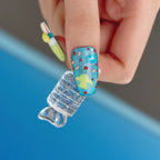 Close-up of a hand holding a 3D press-on coffin nail from the "Candy Night" collection, featuring a blue glitter base with yellow and light blue 3D shapes, red dots, and pink accents. Another nail in the background displays a colorful design, emphasizing the playful, candy-inspired theme of the product.