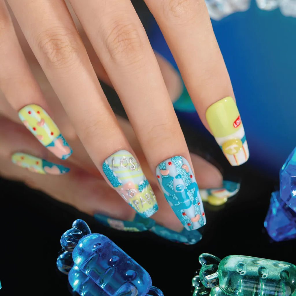 Close-up of a hand wearing 3D press-on nails from the "Candy Night" collection—coffin-shaped (medium length) with vibrant, playful designs: yellow nails with red dots and blue accents, a nail with "LOS" text and glitter, a blue-themed nail with candy motifs, and a Lotion bottle-inspired nail. The background includes translucent blue and green candy-shaped objects, emphasizing the sweet, whimsical theme.