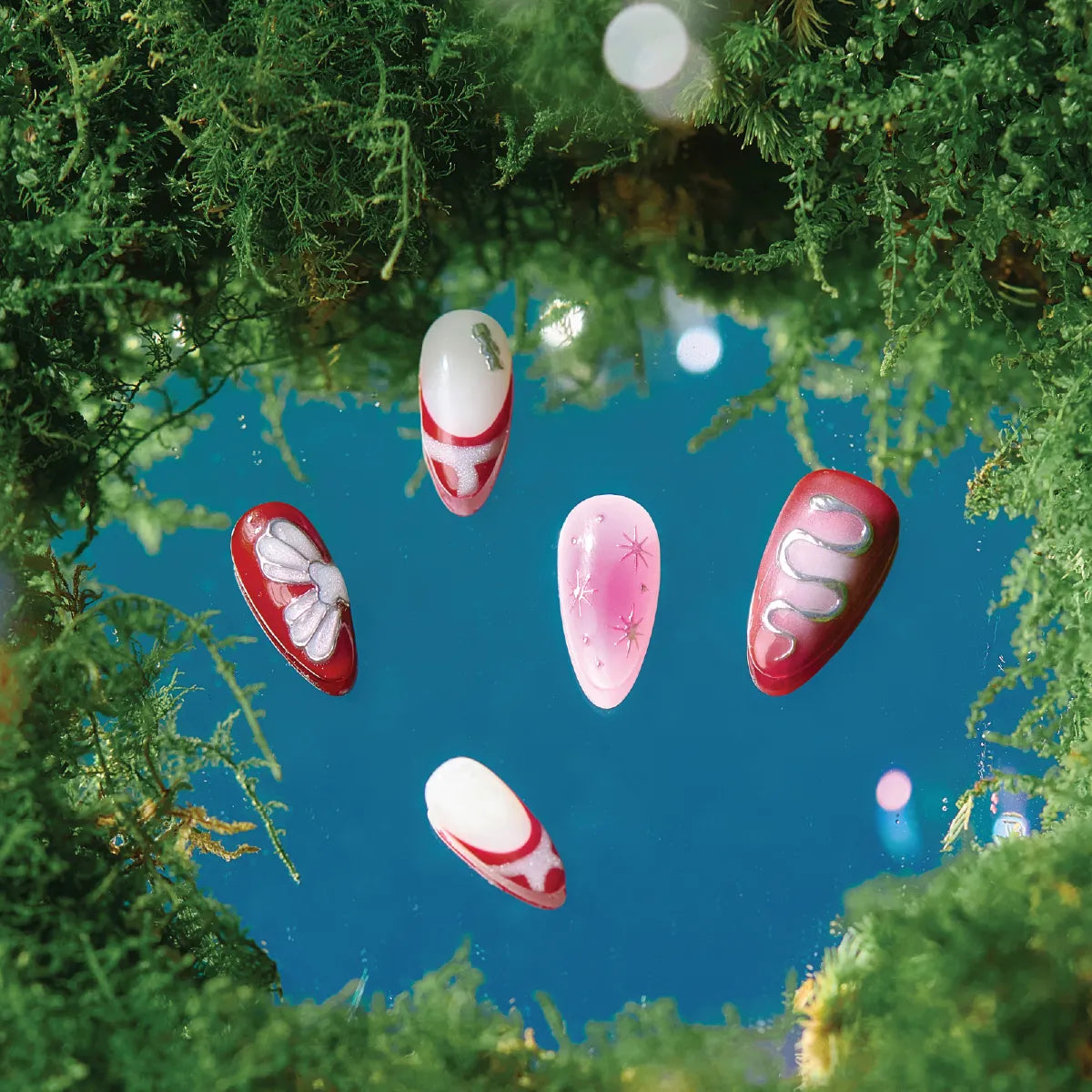 Five medium-length almond-shaped false nails from the Park Select "Cherry Stardust" collection, featuring vibrant designs including red with white bows, pink with star accents, red with silver snake patterns, and red-and-white geometric motifs, displayed on a blue surface surrounded by lush green moss.