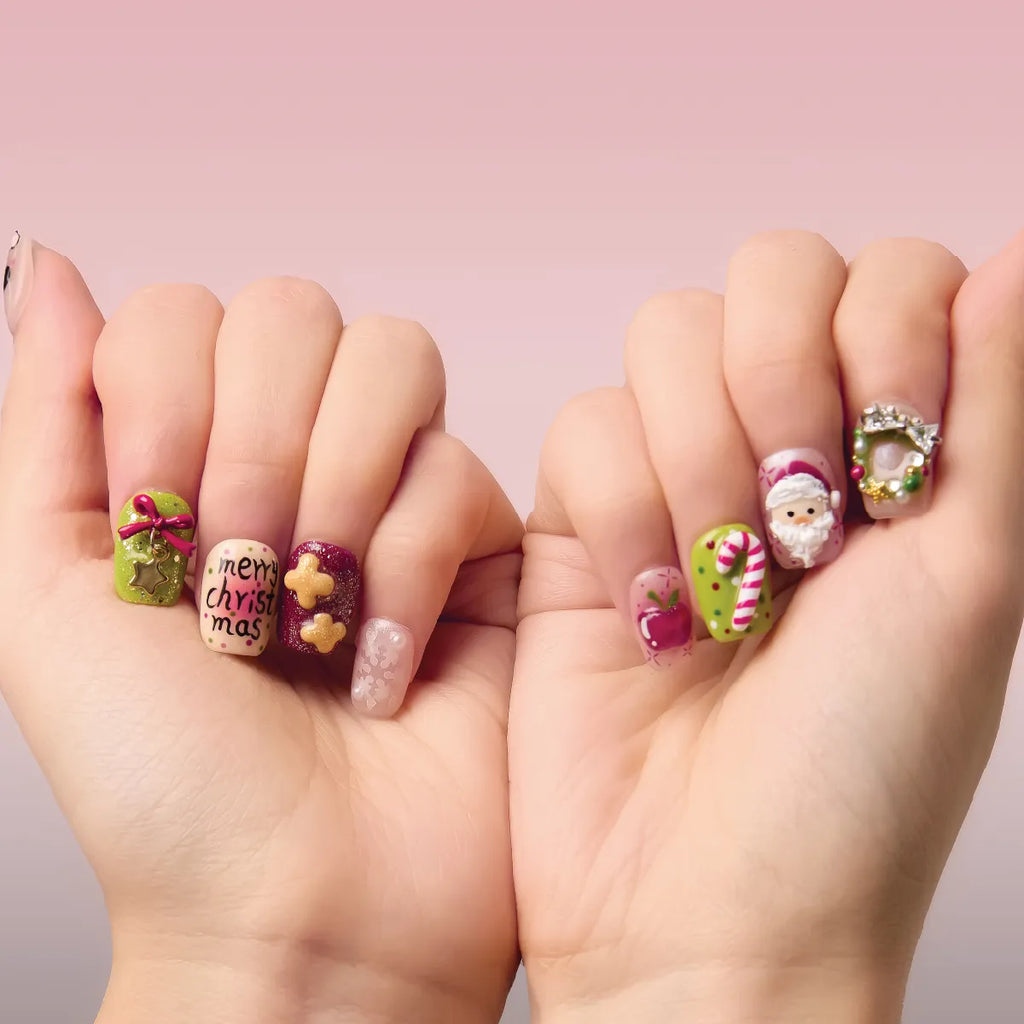 Close-up of two hands holding Christmas Sonata 3D square short press-on nails, showcasing festive designs: "merry christmas" text, bows, stars, gingerbread, Santa, candy canes, and wreaths, against a soft pink background.