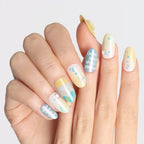 A hand displays medium-length, round 3D press-on nails from the Blossom Mirror collection. The nails feature a soft pastel color palette (yellows, blues, whites) with celestial motifs (moons, stars), text, and decorative accents (pearls, dots), creating a whimsical, dreamy aesthetic.