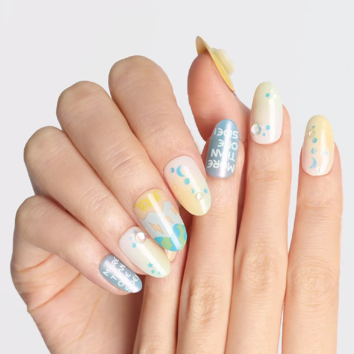 A hand displays medium-length, round 3D press-on nails from the Blossom Mirror collection. The nails feature a soft pastel color palette (yellows, blues, whites) with celestial motifs (moons, stars), text, and decorative accents (pearls, dots), creating a whimsical, dreamy aesthetic.
