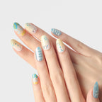 Close-up of hands wearing Blossom Mirror 3D press-on nails (medium length, round shape) with vibrant, whimsical designs: pastel yellow-to-white gradients, blue bases with white text, geometric patterns, celestial motifs (stars, moons, dots), and rhinestone accents, all with a glossy finish.
