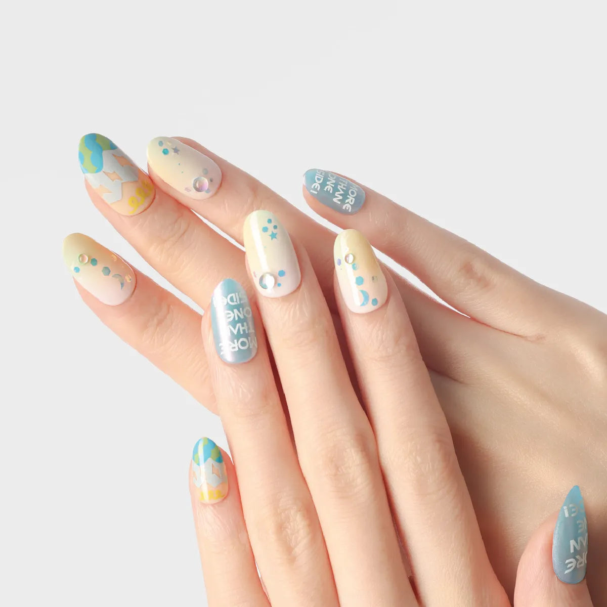 Close-up of hands wearing Blossom Mirror 3D press-on nails (medium length, round shape) with vibrant, whimsical designs: pastel yellow-to-white gradients, blue bases with white text, geometric patterns, celestial motifs (stars, moons, dots), and rhinestone accents, all with a glossy finish.
