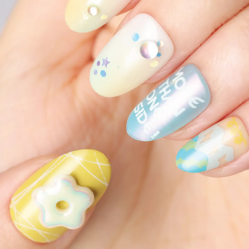 Close-up of a hand wearing Blossom Mirror 3D press-on nails (round shape) with pastel designs: thumb has a yellow grid base with a 3D star charm; index features a pale yellow base with stars, dots, and a pearl; middle nail has an iridescent light blue base with white text; ring nail includes a 3D figure on a pale blue base; pinky has a pale yellow base with a pearl and dot. Designs incorporate soft pastels, 3D embellishments, and playful details.
