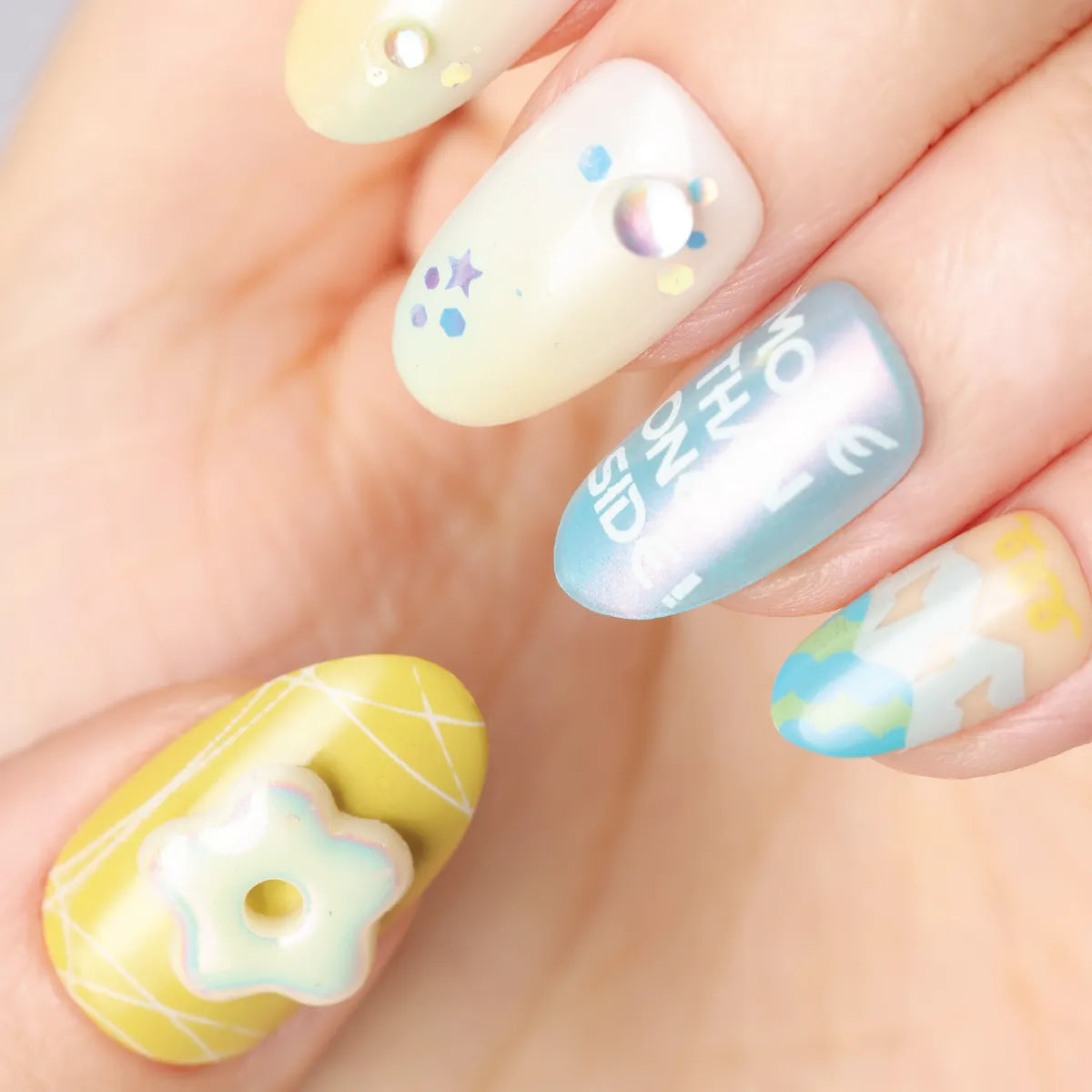 Close-up of a hand wearing Blossom Mirror 3D press-on nails (round shape) with pastel designs: thumb has a yellow grid base with a 3D star charm; index features a pale yellow base with stars, dots, and a pearl; middle nail has an iridescent light blue base with white text; ring nail includes a 3D figure on a pale blue base; pinky has a pale yellow base with a pearl and dot. Designs incorporate soft pastels, 3D embellishments, and playful details.