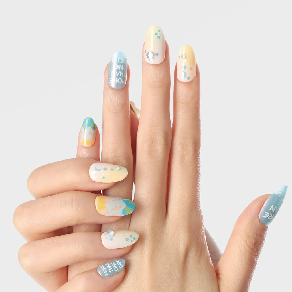 Close-up of a hand displaying 3D press-on nails from the Blossom Mirror collection, featuring round, medium-length nails with pastel blue, yellow, and white bases adorned with stars, dots, text, and geometric patterns.