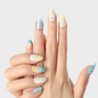 Close-up of a hand displaying 3D press-on nails from the Blossom Mirror collection, featuring round, medium-length nails with pastel blue, yellow, and white bases adorned with stars, dots, text, and geometric patterns.