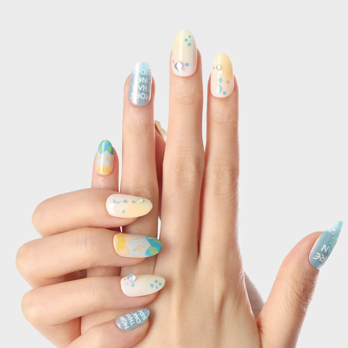Close-up of a hand displaying 3D press-on nails from the Blossom Mirror collection, featuring round, medium-length nails with pastel blue, yellow, and white bases adorned with stars, dots, text, and geometric patterns.