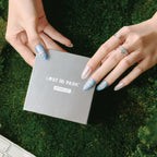 Two hands with blue almond-shaped false nails (featuring delicate patterns) hold a gray "LOST in PARK ARTISAN SET" box, set against a green moss background. The right hand wears silver rings (including a pearl and heart design). The nails are part of the "Blue Frequency" collection from "Park Select," with an almond shape.