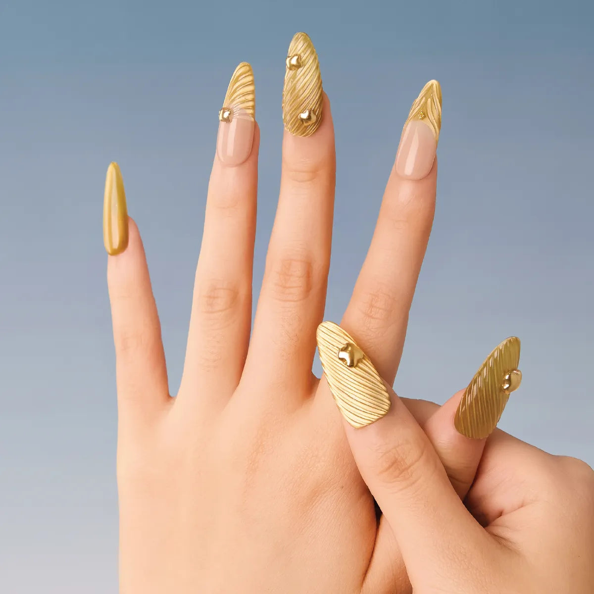 A hand displays Chapter Gold 3D press-on nails (almond shape, long length) with a gold, textured (striped) design and small gold accents, showcasing a luxurious look against a blue background.