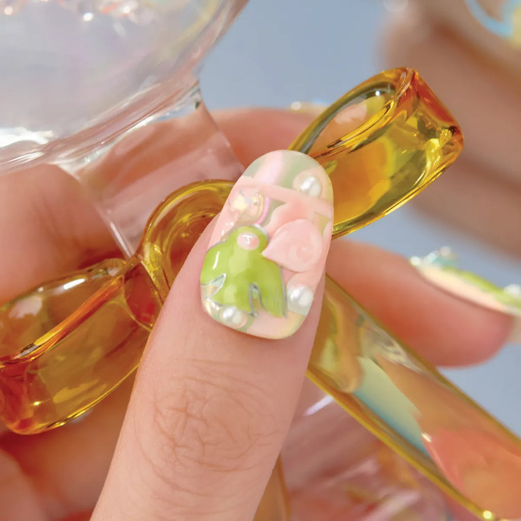 Close-up of a hand holding yellow sunglasses, showcasing a 3D press-on nail from the Bubblegum Heart collection. The round, medium-length nail has a pastel pink base with a green frog, pink bow, and pearl accents, featuring a playful, whimsical design.