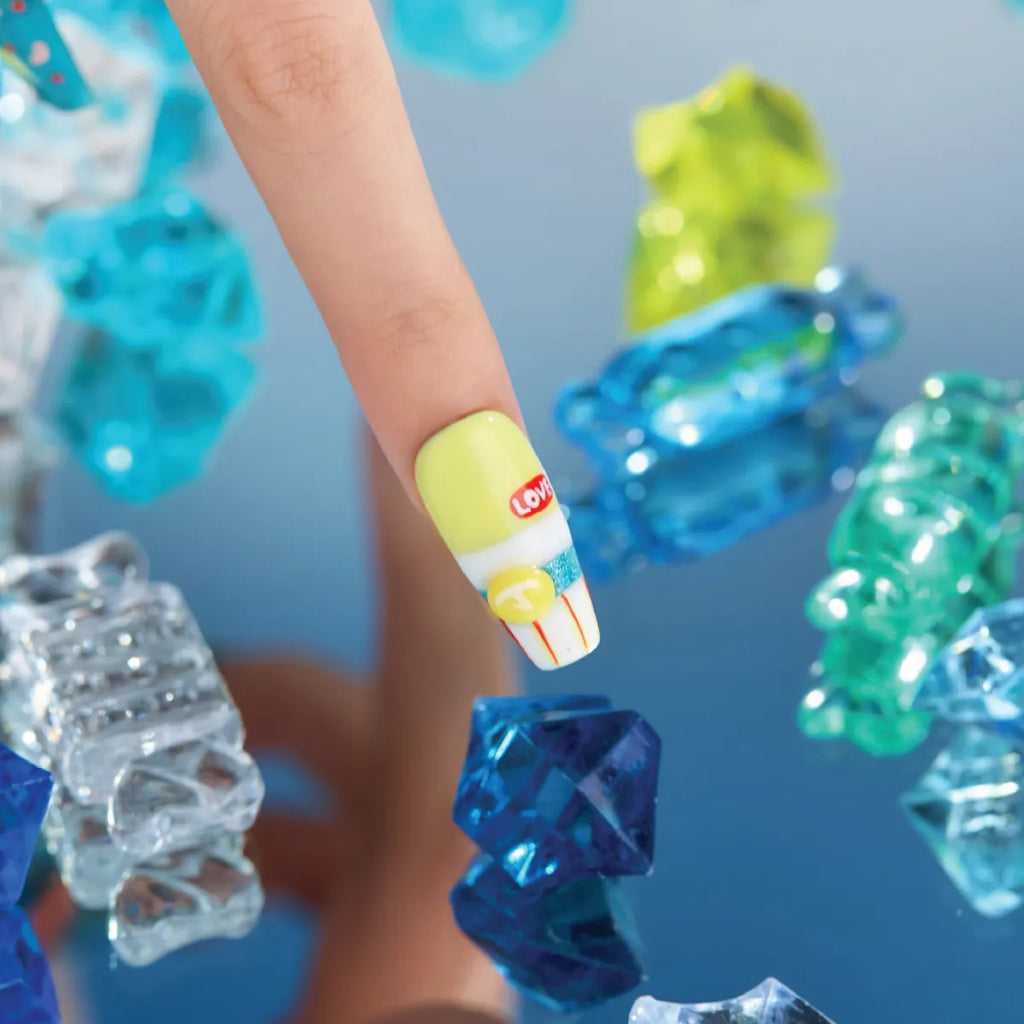 A hand showcases a medium-length, coffin-shaped 3D press-on nail from the "Candy Night" collection, adorned with a yellow base, white stripe, red "LOVE" text, blue glitter stripe, yellow "T" button, and red vertical lines. The nail is positioned over colorful, translucent 3D candy-like decorations (blue, green, clear) on a blue surface.