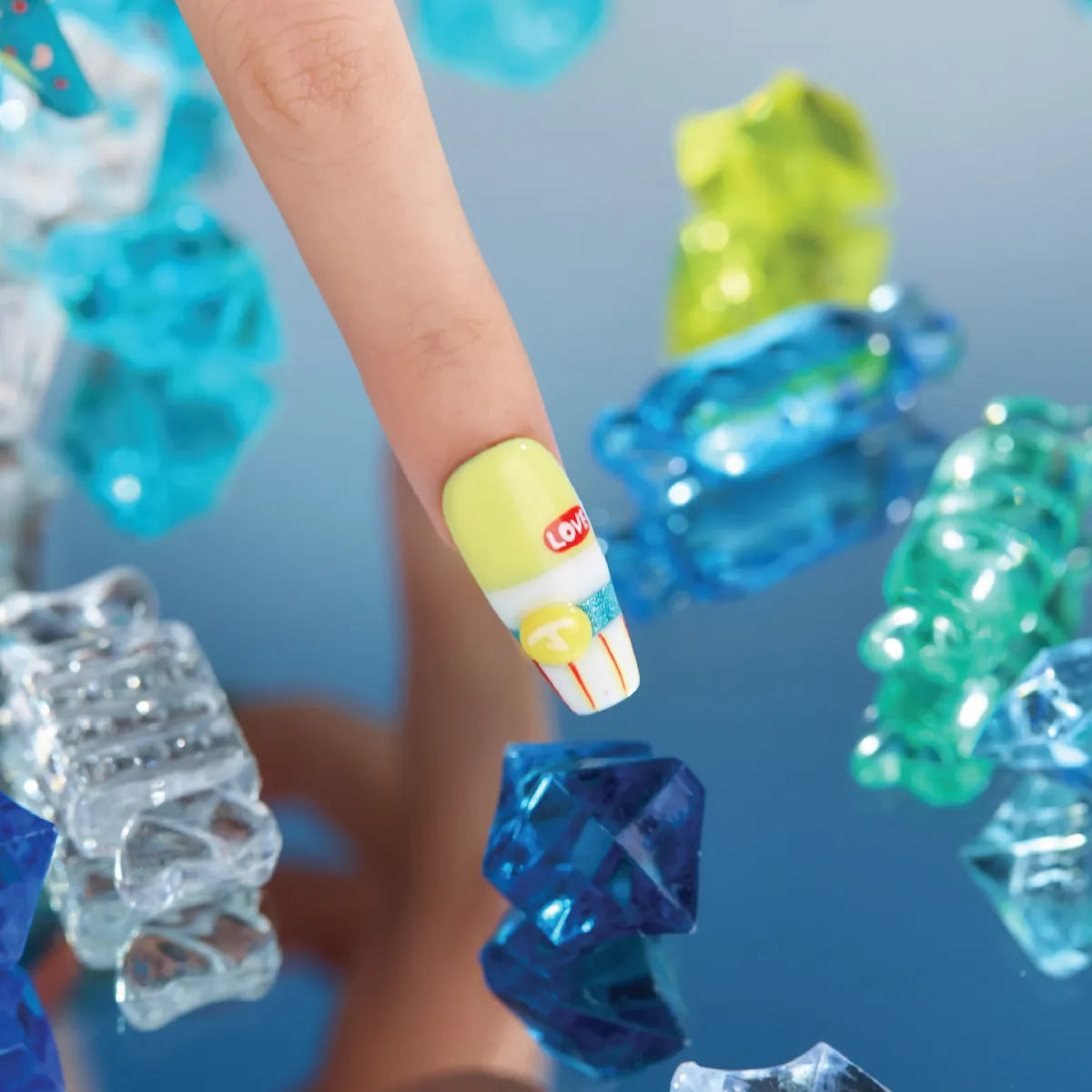A hand showcases a medium-length, coffin-shaped 3D press-on nail from the "Candy Night" collection, adorned with a yellow base, white stripe, red "LOVE" text, blue glitter stripe, yellow "T" button, and red vertical lines. The nail is positioned over colorful, translucent 3D candy-like decorations (blue, green, clear) on a blue surface.