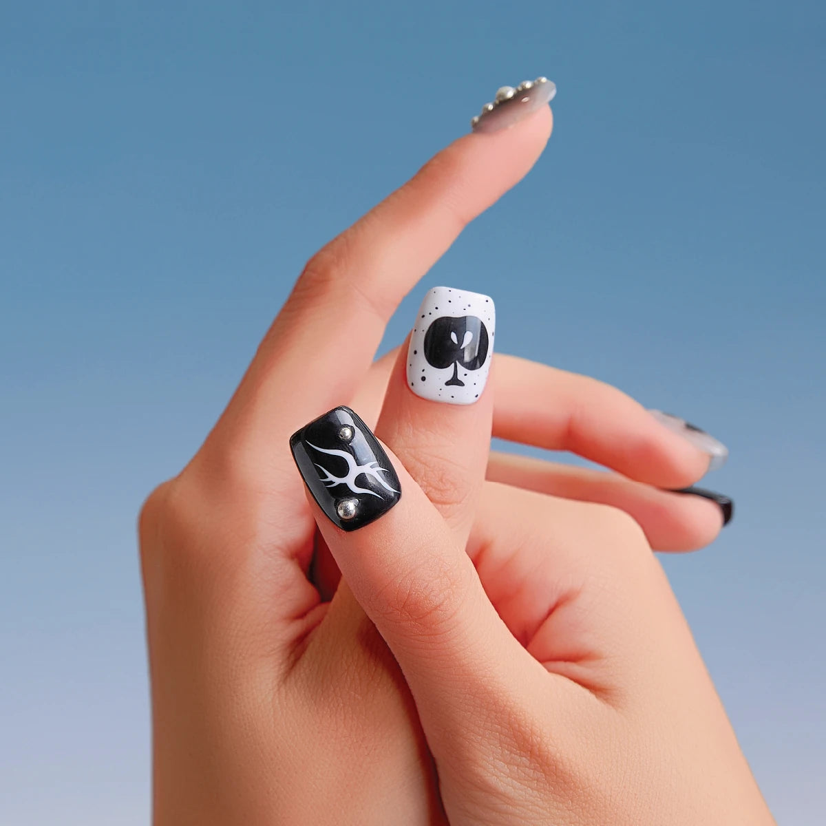 Close-up of two hands showcasing Astro Courier 3D press-on nails (short, square shape). Designs include a black nail with white flame-like patterns and two silver rhinestones, a white nail with black polka dots and a black club symbol, and a clear thumb nail with three silver rhinestones, set against a blue gradient background.
