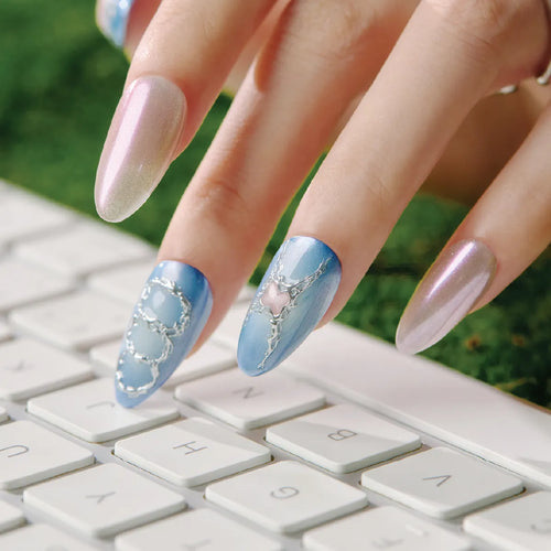 Close-up of hands with almond-shaped false nails from the "Blue Frequency" Park Select collection: two nails feature blue bases with silver decorative designs (one with circular motifs, one with a heart and branch-like pattern), while the other two have metallic pinkish-purple finishes. Hands rest on a white keyboard, with a green background (likely grass) visible. Nails are medium length, designed for easy application.