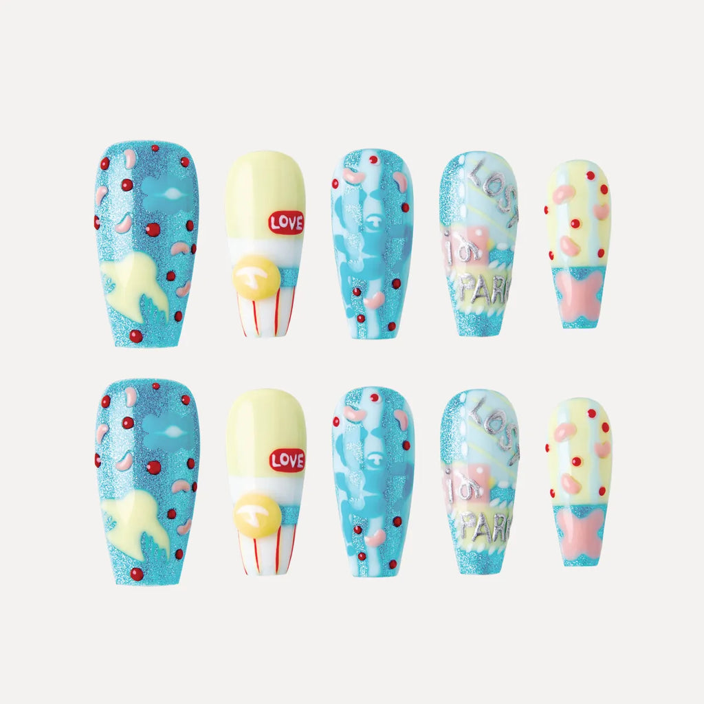Image of 3D press-on nails from the "Candy Night" collection, featuring coffin-shaped, medium-length nails with vibrant, playful designs. Two rows of five nails each showcase colorful patterns: blue glitter bases with yellow stars and red/pink polka dots; yellow-and-white nails with "LOVE" text and a yellow circular accent; blue nails with abstract pink shapes and red dots; nails with "Los for Paris" text in silver on a blue/pink background; and yellow nails with pink shapes and blue glitter accents.