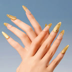 Chapter Gold 3D press-on nails (almond shape, long length) with gold textured designs and small decorative accents, displayed on a hand against a blue background.