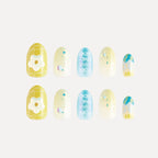 Blossom Mirror 3D press-on nails (round, medium length) featuring designs: yellow base with white flower/grid, light yellow with colorful dots/balls, blue with reversed text, and blue-yellow gradient with pattern. Two matching sets displayed.