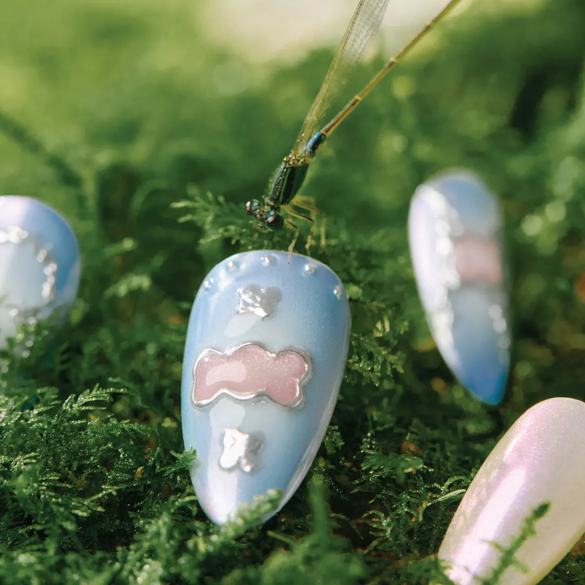 Close-up of almond-shaped "Blue Frequency" false nails (from the Park Select collection) featuring a light blue base, pink bow, and silver accents (dots, shapes), placed on vibrant green moss with a dragonfly perched on one nail, creating a whimsical, nature-inspired scene.