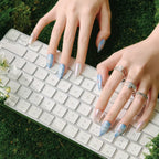 Hands with almond-shaped false nails (blue with silver accents, pearl-like tips) typing on a white keyboard, adorned with silver rings, set against a mossy green background. Product: Blue Frequency (Park Select collection).