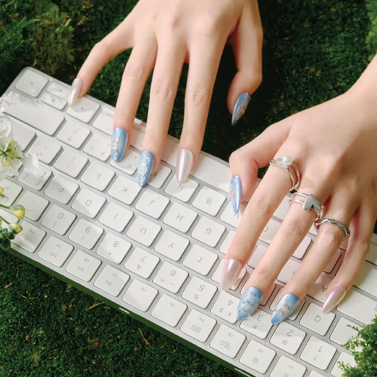 Hands with almond-shaped false nails (blue with silver accents, pearl-like tips) typing on a white keyboard, adorned with silver rings, set against a mossy green background. Product: Blue Frequency (Park Select collection).