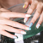 Close-up of two hands (left and right) resting on a glass surface with water droplets, showcasing 3D round, medium-length press-on nails from the Blossom Mirror collection. Nails feature pastel hues (white, yellow, blue, gray) with designs including the text “MORE THAN ONE SIDE,” celestial motifs (moons, stars), rhinestones, and abstract patterns, highlighting the detailed, artistic nail art.