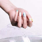 Close-up of a hand with medium-length, round 3D press-on nails from the Blossom Mirror collection, featuring pastel-colored designs (stars, text, and small decorative elements) on each nail. The hand rests on a transparent surface with water droplets, highlighting the nails’ glossy finish and intricate details.