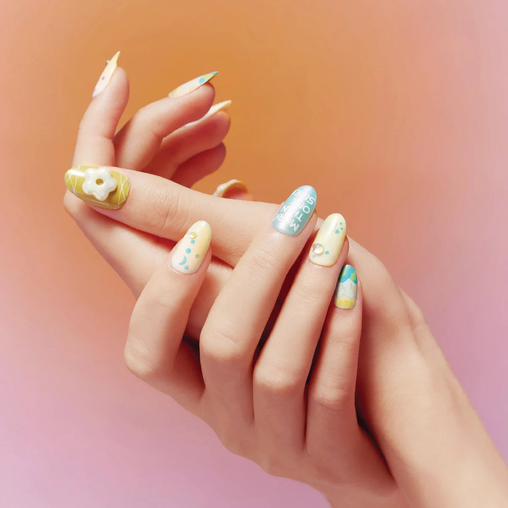 Two hands showcase Blossom Mirror 3D Press on Nails (round shape) with whimsical, pastel designs: a 3D flower on a yellow base, a light blue nail with text, and others adorned with dots, stars, and pearl accents. The background features a warm gradient from orange to pink, emphasizing the playful, detailed nail art.
