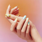 Two hands showcase Blossom Mirror 3D Press on Nails (round shape) with whimsical, pastel designs: a 3D flower on a yellow base, a light blue nail with text, and others adorned with dots, stars, and pearl accents. The background features a warm gradient from orange to pink, emphasizing the playful, detailed nail art.
