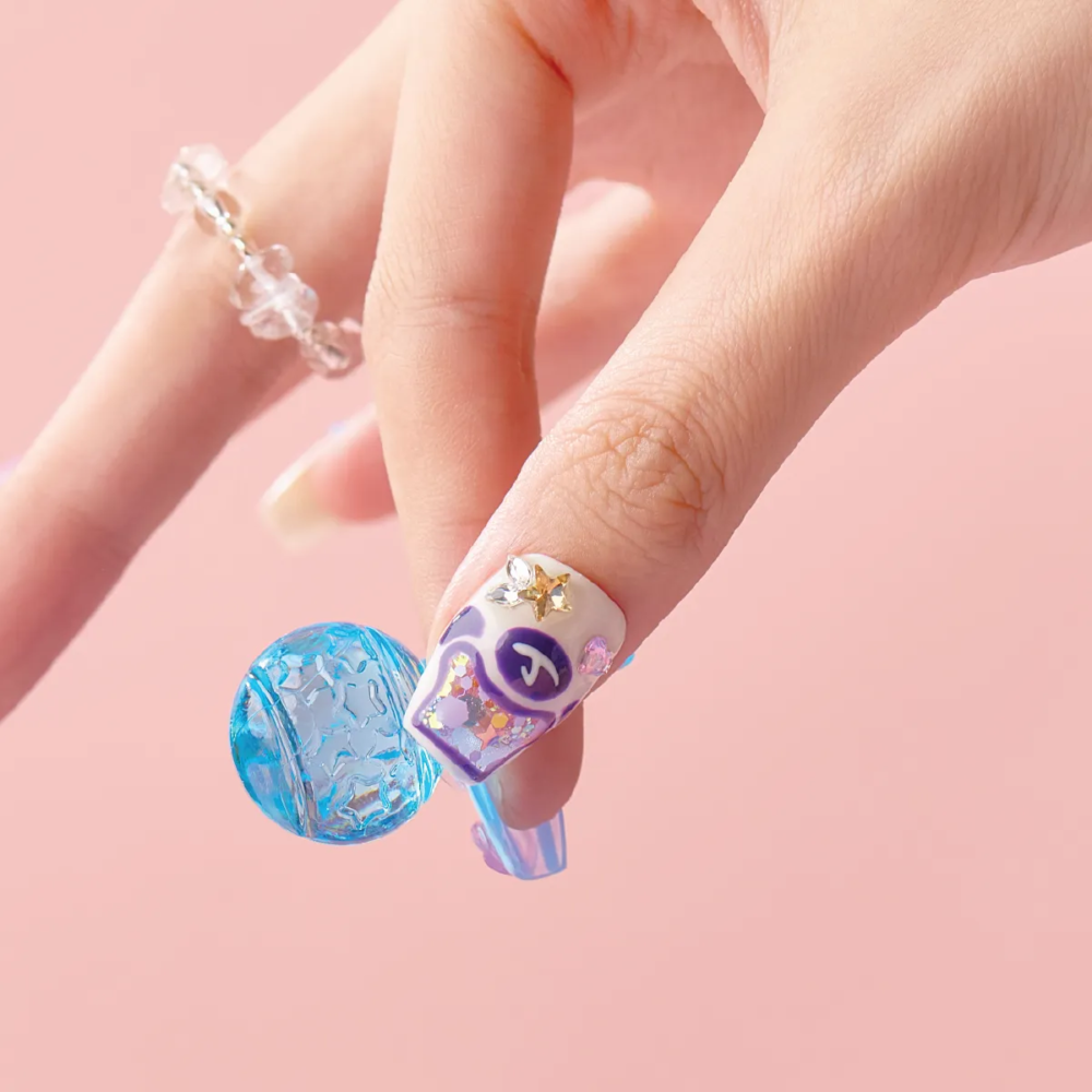 Close-up of a hand wearing a Blue Lollipop press-on nail with purple and white 3D candy details, holding a blue textured bead.