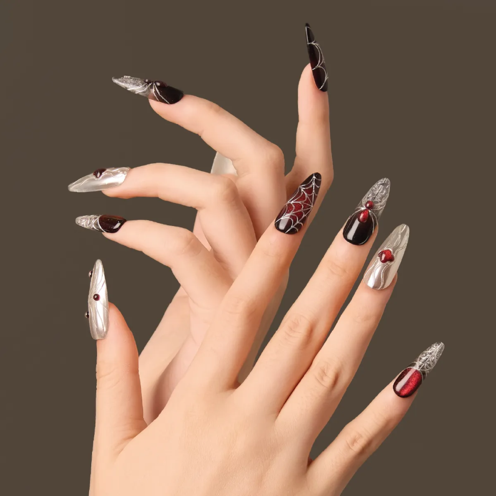 Artistic hand pose showcasing the “Crimson Whisper” nails with metallic silver designs, deep red shimmer, and white spiderweb graphics.