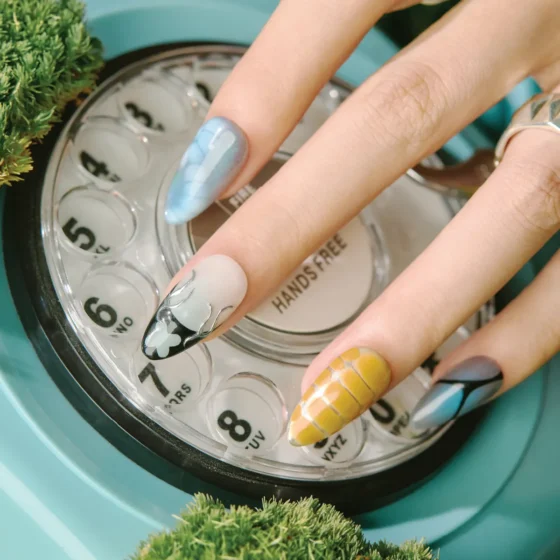 Close-up of abstract blue, yellow grid, and black-and-white flame design nails resting on a rotary phone dial