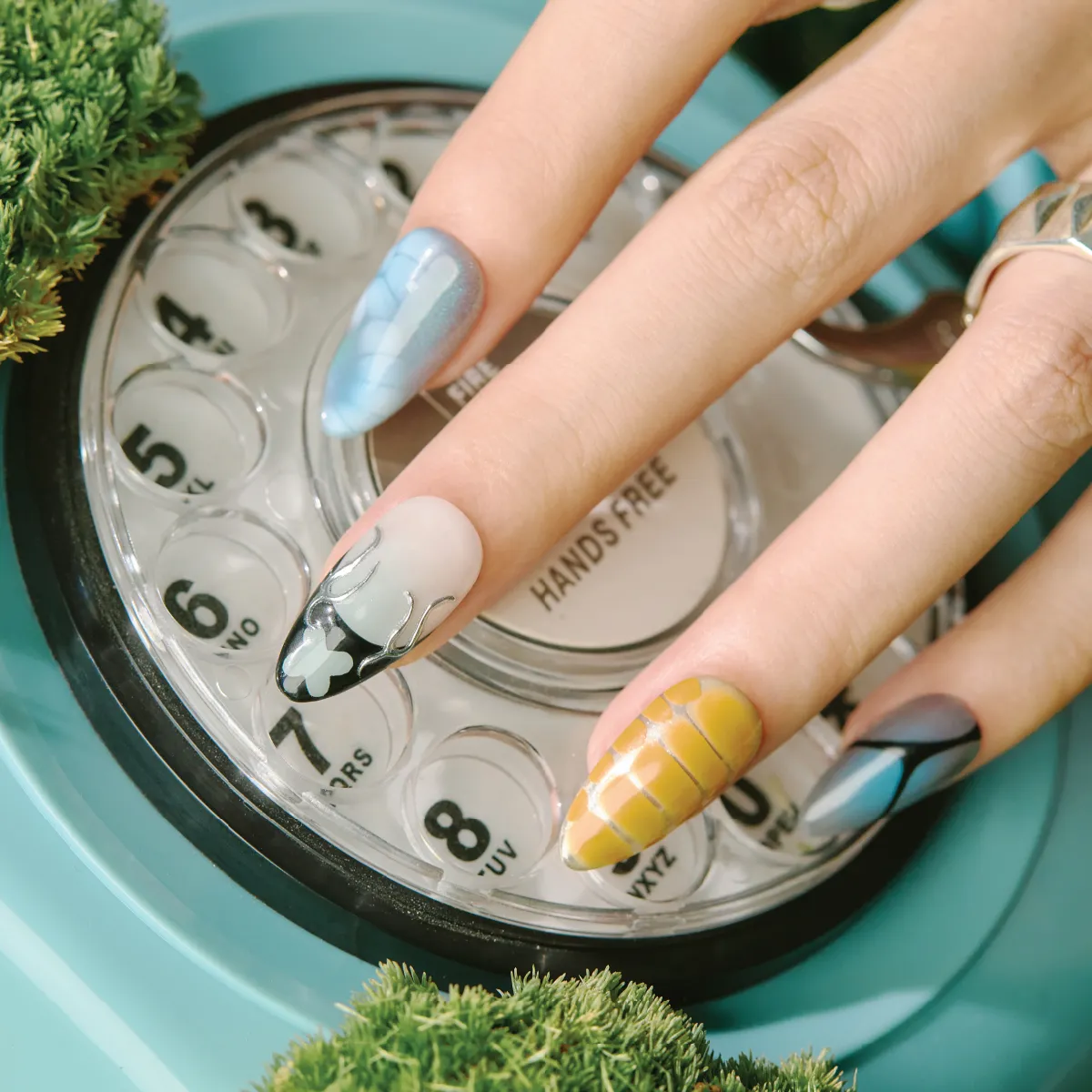 Close-up of abstract blue, yellow grid, and black-and-white flame design nails resting on a rotary phone dial