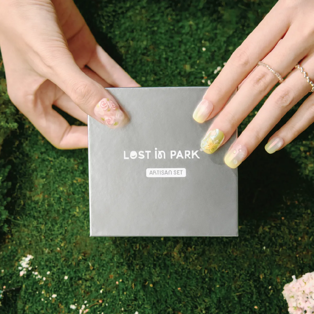 Hands wearing Dewy Garden press-on nails holding a LOST in PARK Artisan Set box over a grass background