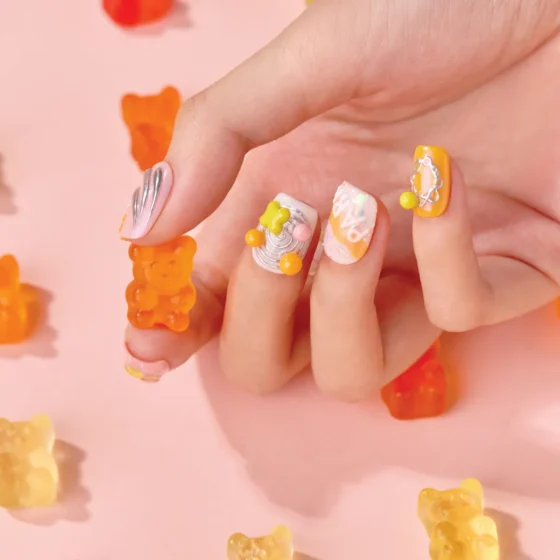 Hand with vibrant orange and pink 3D press-on nails featuring gummy bear designs, holding a translucent gummy bear candy on a pastel pink background.
