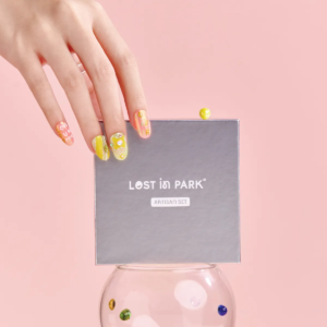 Model hand with bright yellow and orange press-on nails holding a LOST in PARK Artisan Set box.