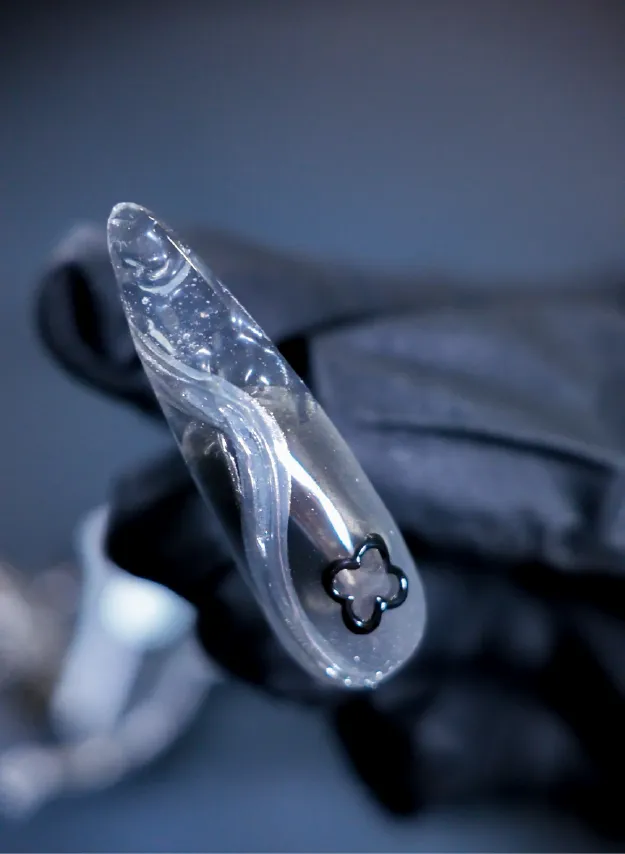 Clear ripple press-on nail with black flower accent held by a gloved hand.