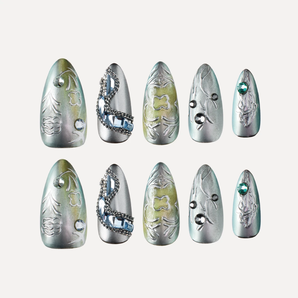 Medium almond press on nails flat lay in silver green tones with embossed patterns chains and gemstone details